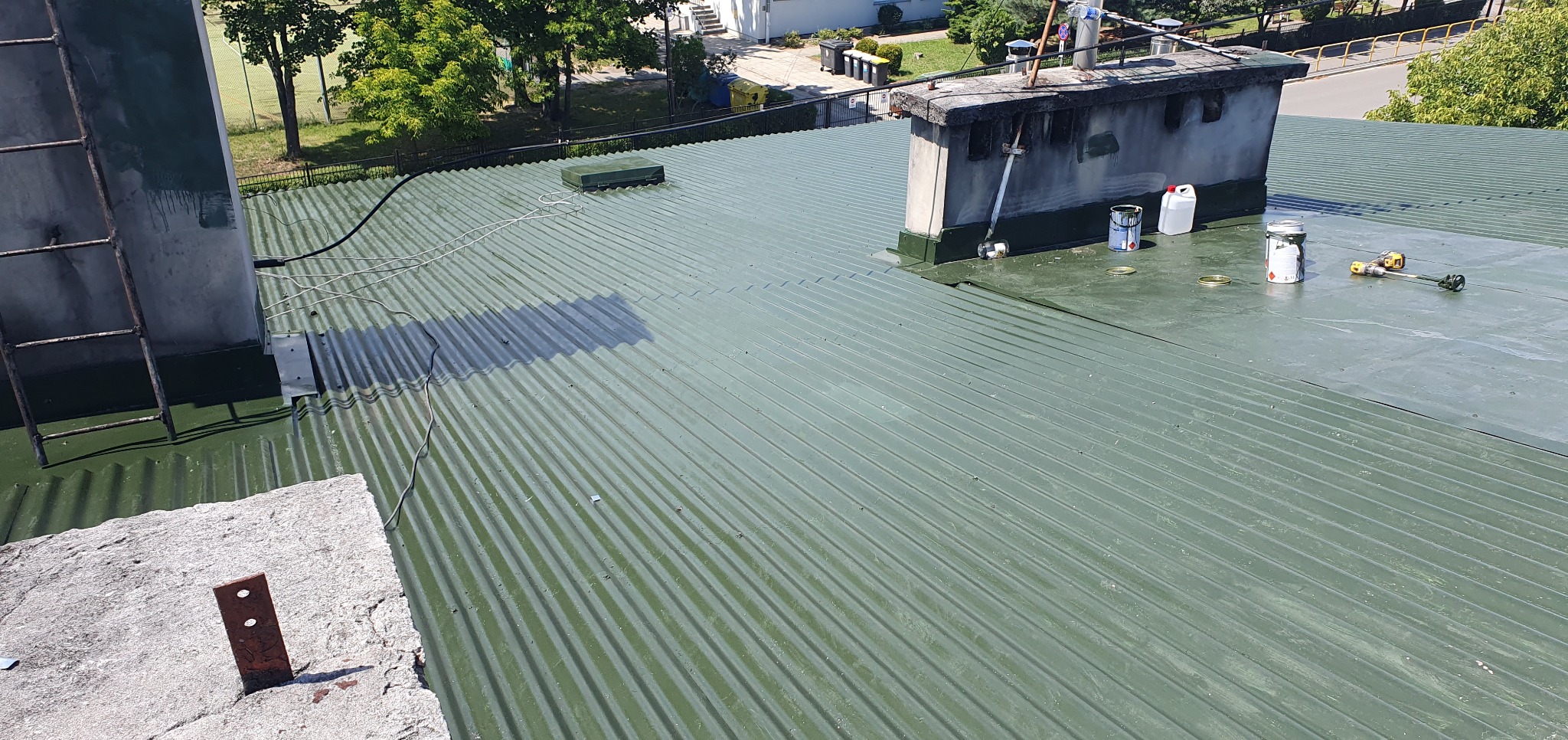 Zielony, pomalowany dach z blachy falistej. Widoczne narzędzia i puszki z farbą. Kominy i drabina na dachu. Ujęcie z góry, w tle zieleń drzew i budynki.