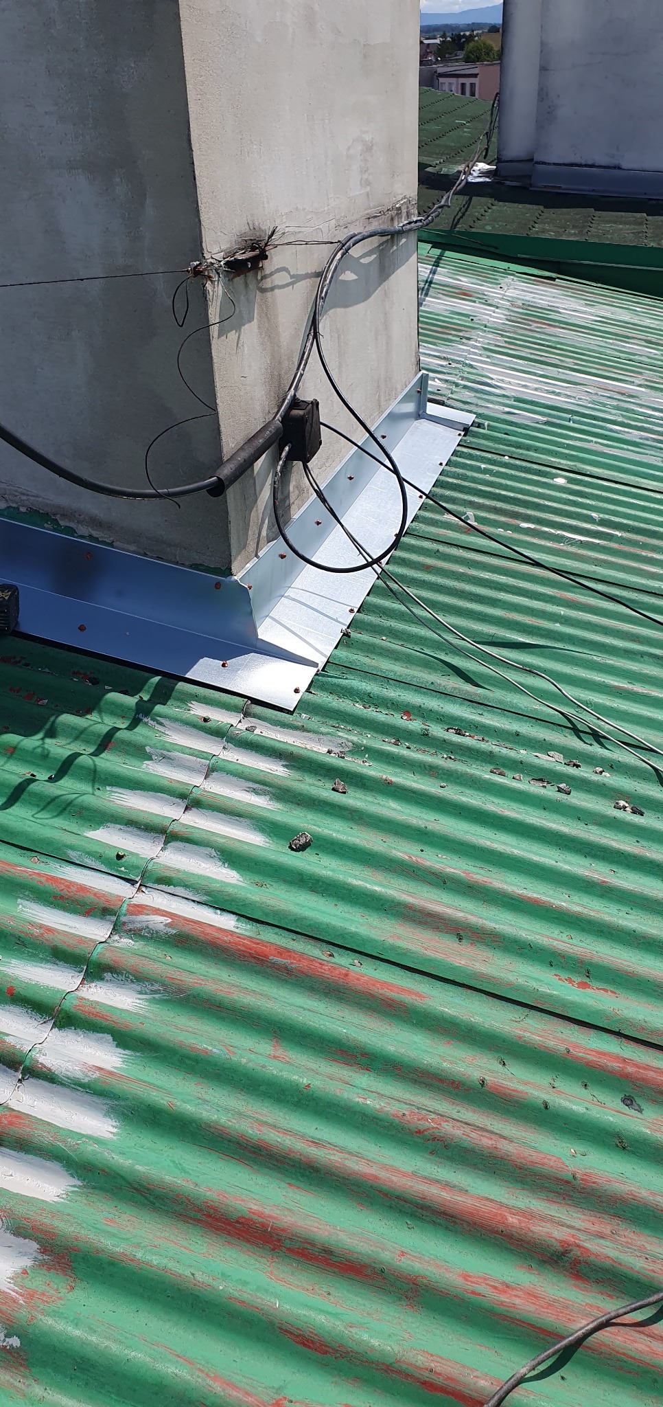 Szczegółowe ujęcie obróbki blacharskiej komina na zielonym, falistym dachu z widocznymi śladami farby. Nowa, srebrna blacha kontrastuje z postarzałym dachem.