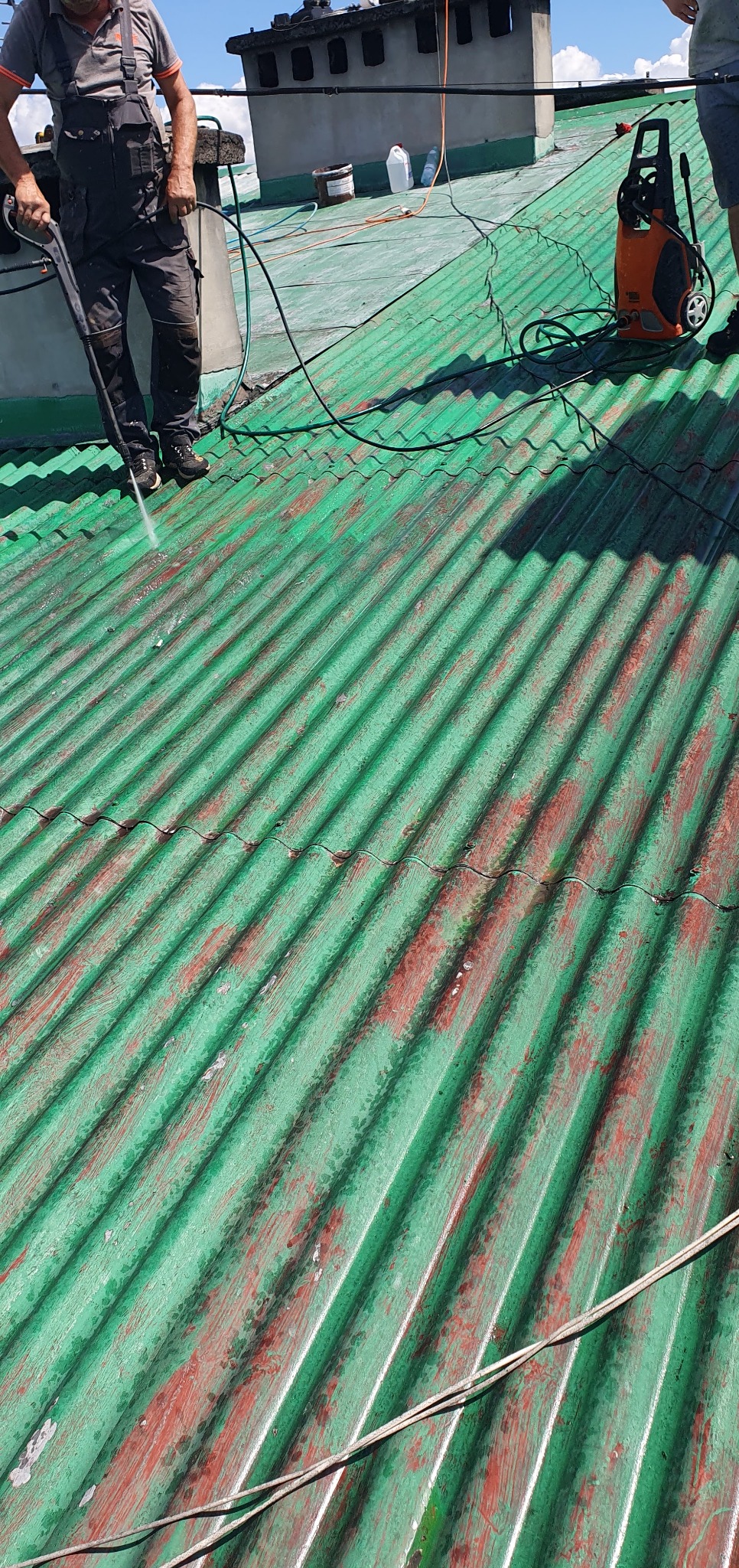 Czyszczenie zielonego, pofalowanego dachu z resztkami czerwonej farby za pomocą myjki ciśnieniowej. Widoczny fragment komina i przewody.