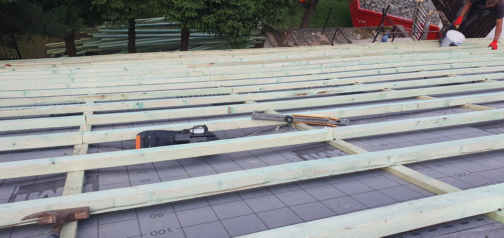 Konstrukcja dachu z łatami, widoczna membrana dachowa, narzędzia: młotek, nóż dekarski, poziomica. W tle drzewa i kontener na gruz.