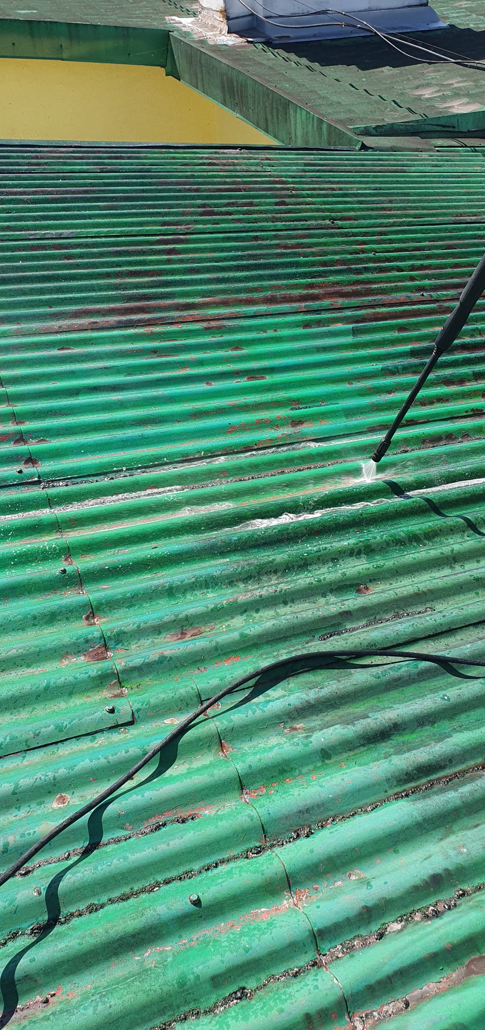 Czyszczenie zielonego, pofalowanego dachu z widocznymi śladami rdzy za pomocą myjki ciśnieniowej. Woda spływa po powierzchni, ukazując odświeżony kolor.