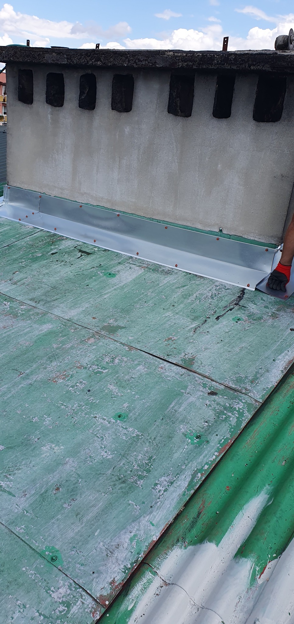 Montaż obróbki blacharskiej komina na zielonym, blaszanym dachu. Widoczne mocowania i fragment dłoni w rękawicy. Komin z szarego betonu z otworami wentylacyjnymi.