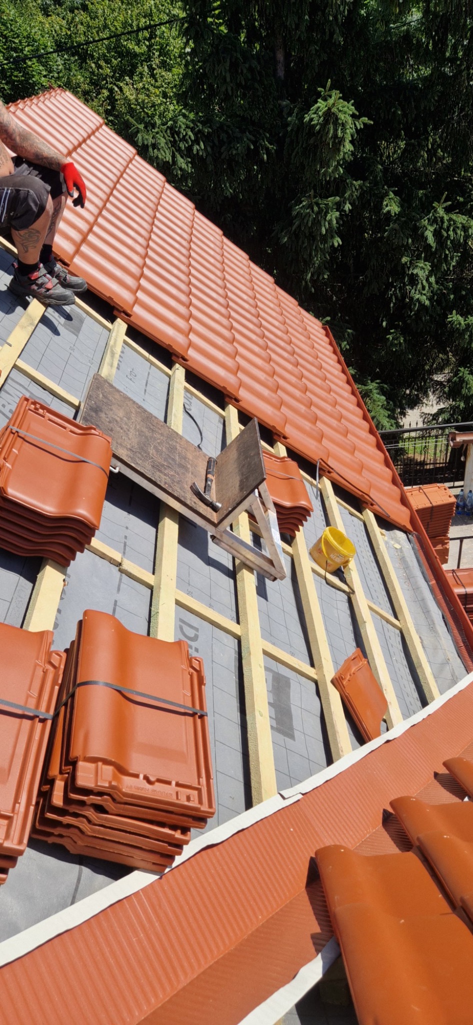 Ceglastoczerwona dachówka karpiówka ułożona na łatach, widoczne narzędzia i fragment dachu w trakcie prac dekarskich na tle zieleni.