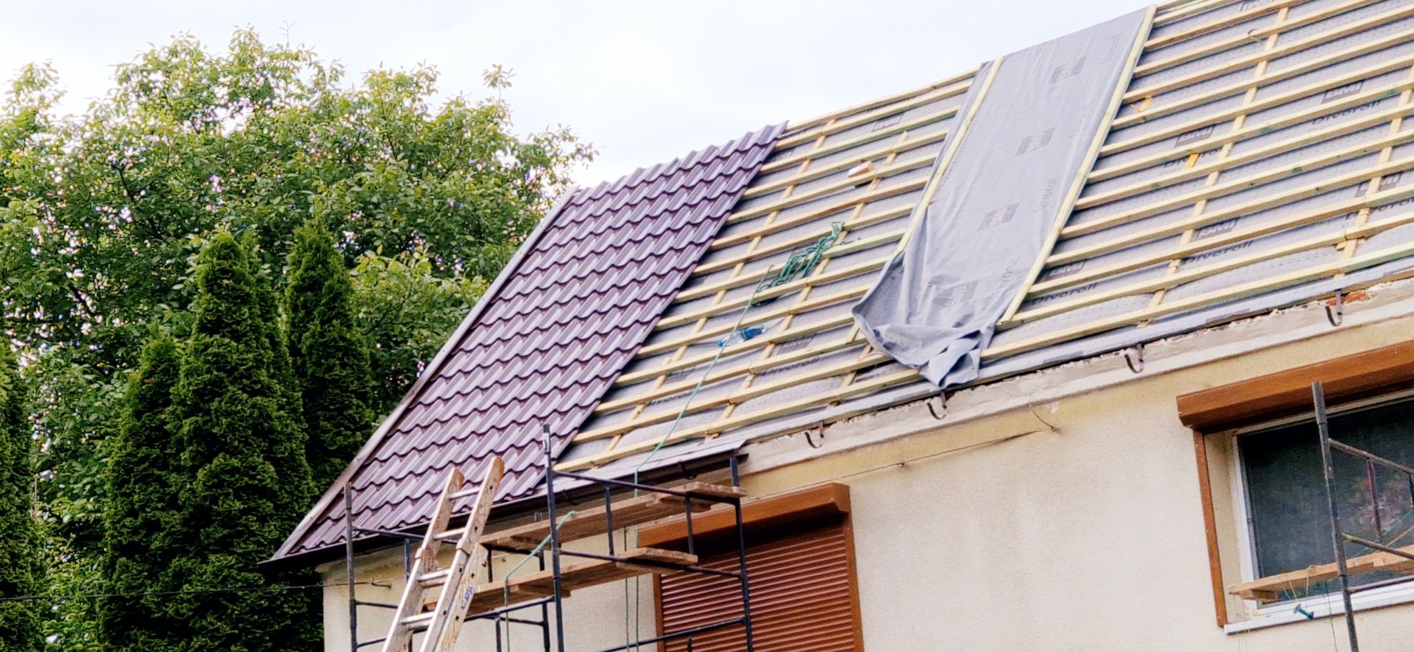Częściowo pokryty dach brązową dachówką, widoczne łaty i membrana. Prace dekarskie w toku, rusztowanie i drabina przy ścianie budynku.