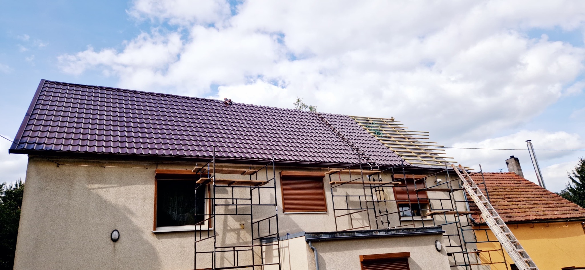 Dom w Nysie z częściowo wymienionym, brązowym dachem z blachodachówki. Widoczne rusztowania i drabina. Część dachu z łatami przed położeniem pokrycia.