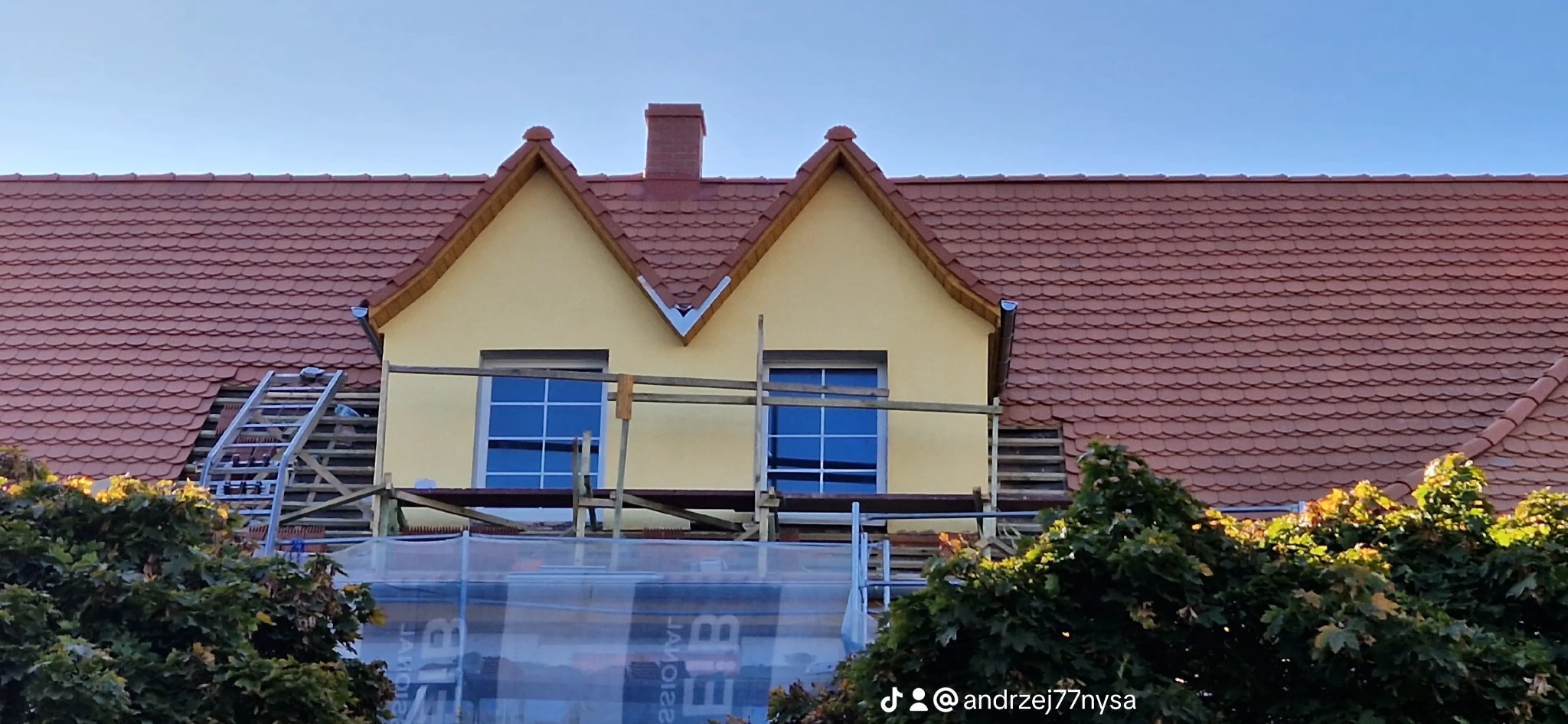 Dom z czerwoną dachówką w trakcie remontu. Widoczne rusztowania, drabina i częściowo zdjęte dachówki. Żółta elewacja i fragmenty zieleni w dolnej części kadru.
