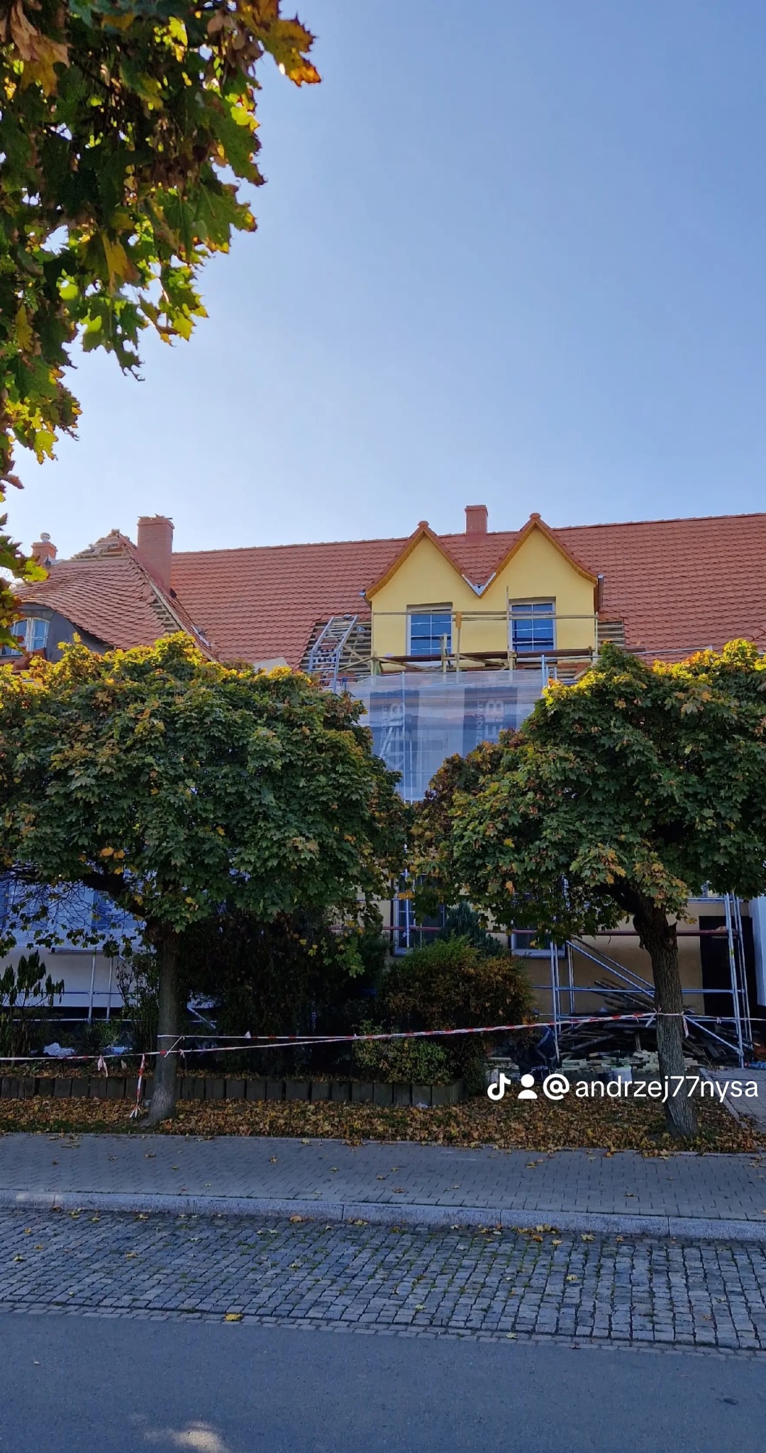 Dom z czerwonym dachem w trakcie remontu, widoczne rusztowania i częściowo zdjęte dachówki. Dwa drzewa na pierwszym planie.