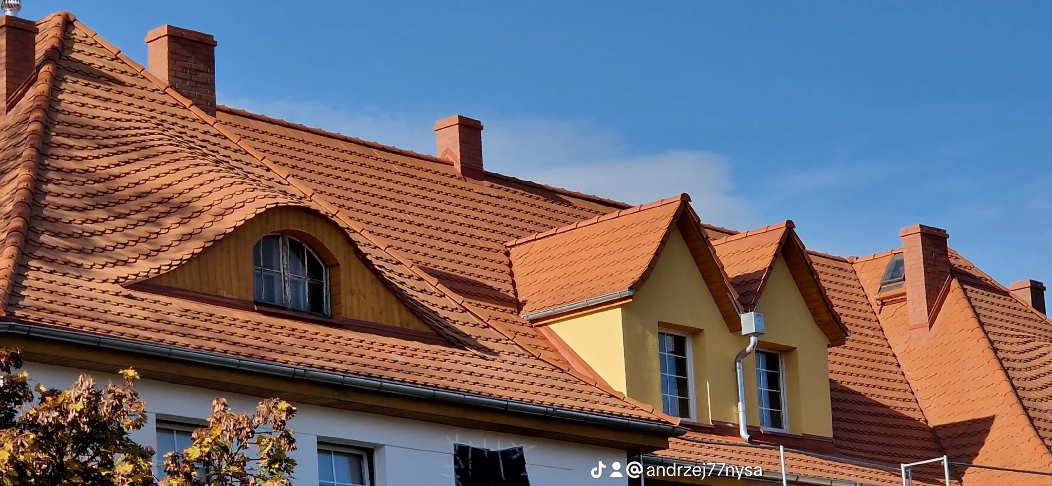 Czerwony dach z dachówki karpiówki na budynku z żółtymi lukarnami i ceglanym kominem, na tle błękitnego nieba. Widoczne rynny i fragment drzewa.