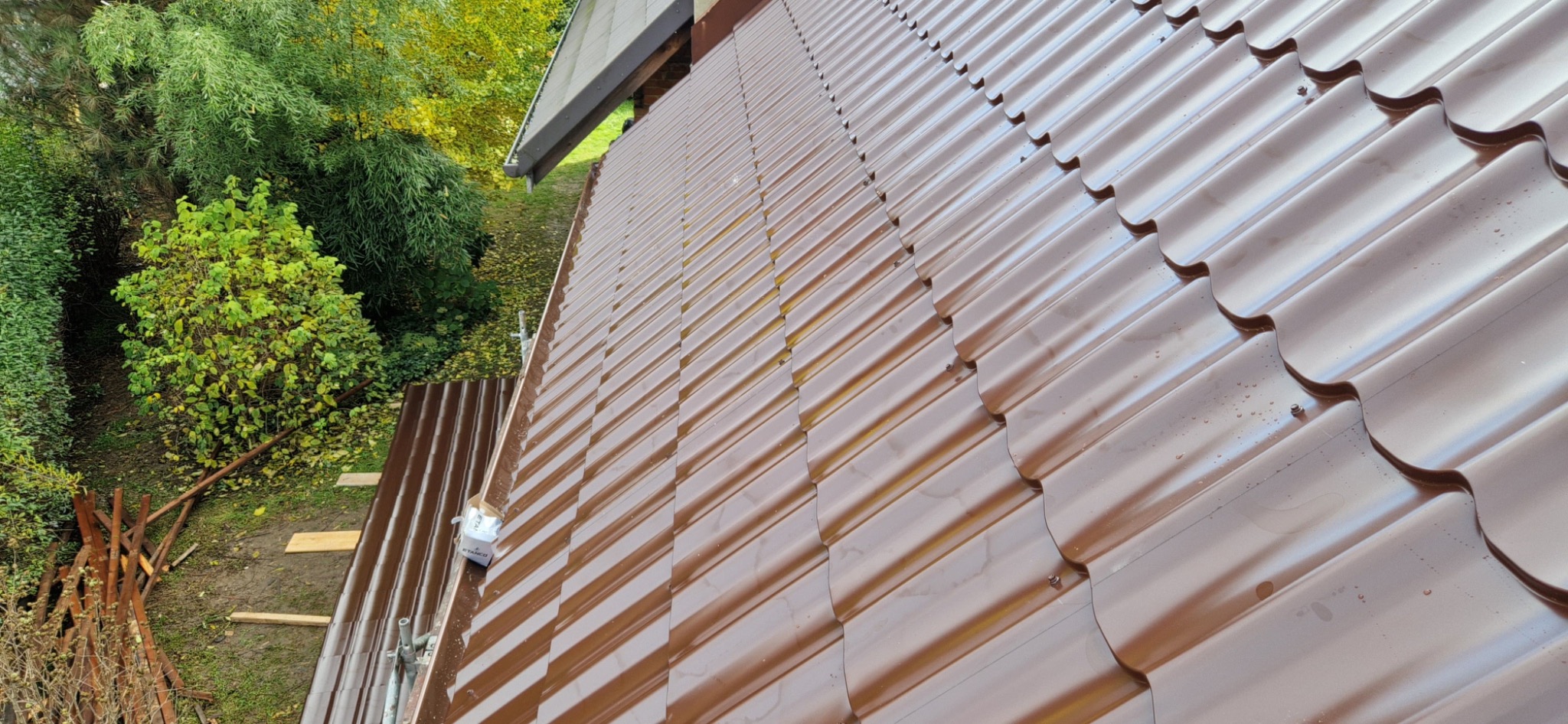 Błyszczące, brązowe dachówki ceramiczne na skośnym dachu domu, widoczne z bliska, z zielenią drzew w tle. Nowe pokrycie dachowe w słoneczny dzień.