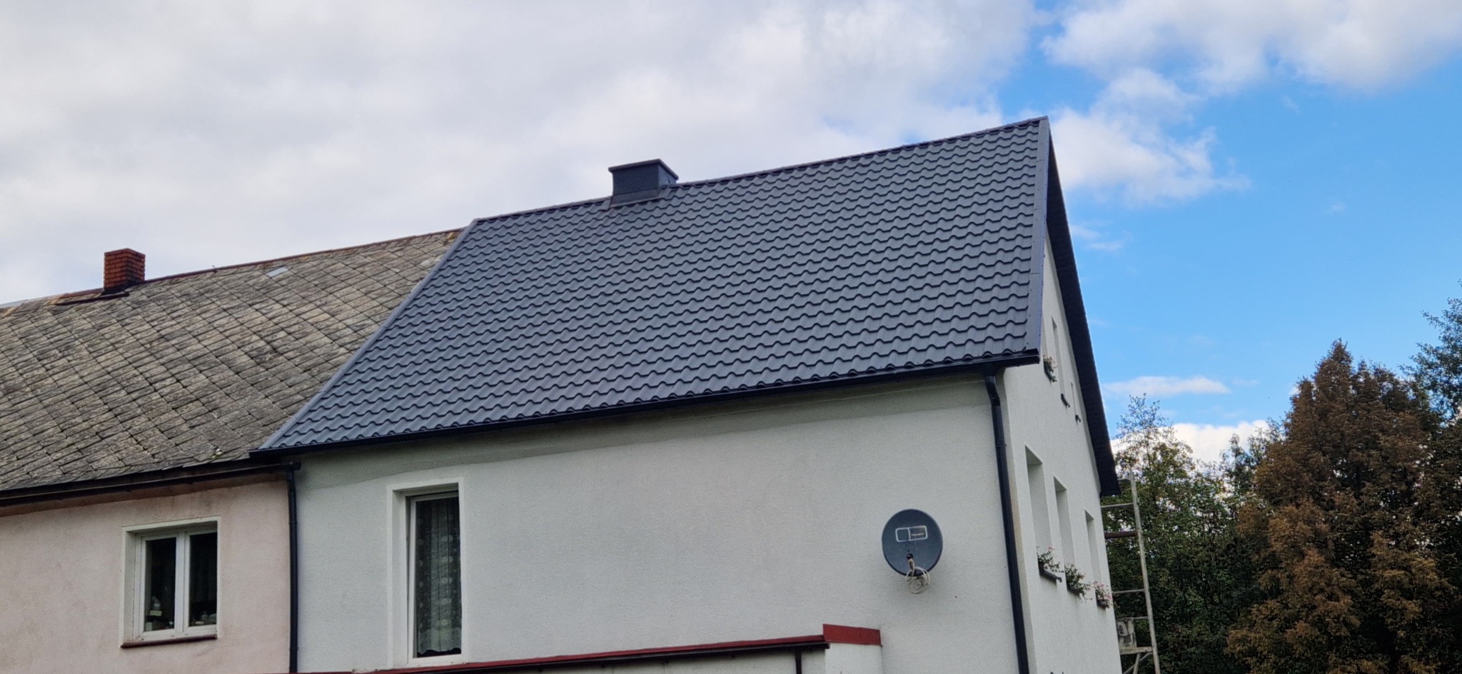 Dom z nowym, ciemnoszarym dachem z blachodachówki trapezowej, kontrastującym z białą elewacją. Widoczny fragment starego dachu sąsiedniego budynku.