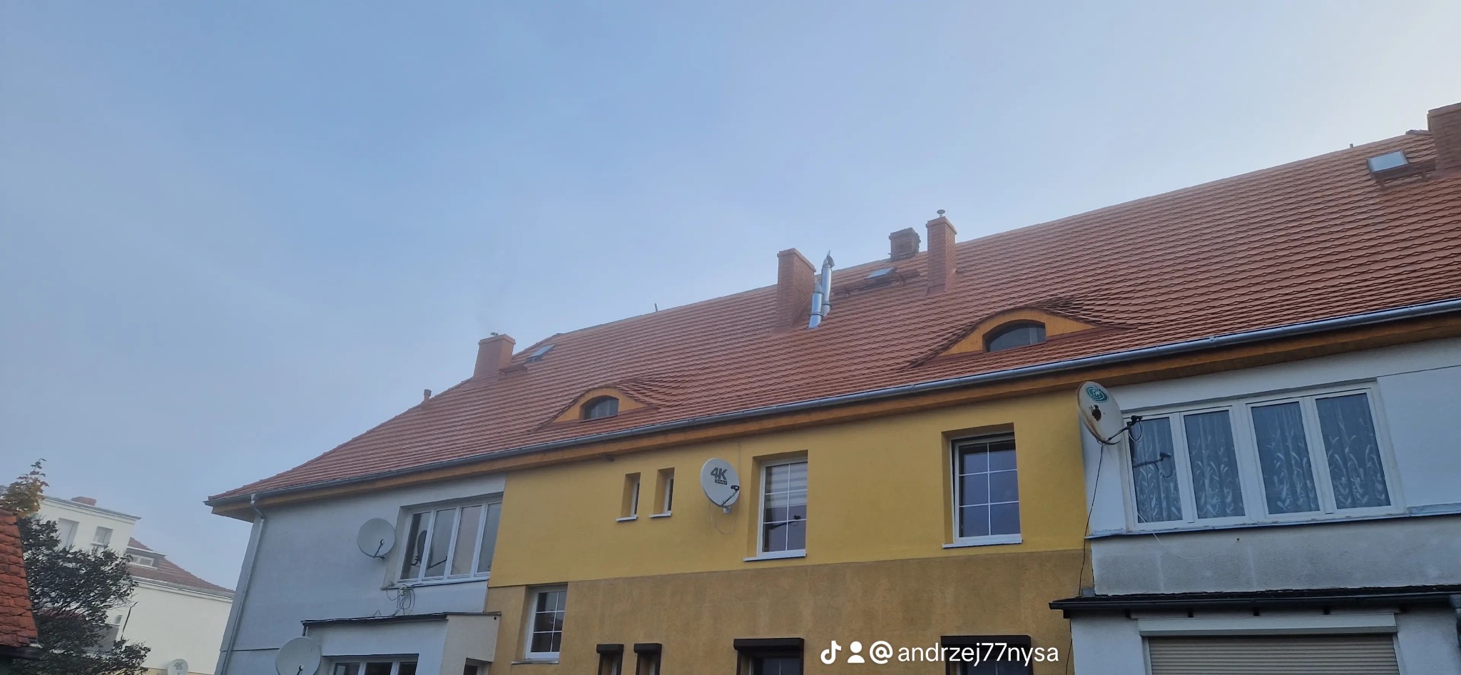 Czerwony dach z oknami typu wole oko na żółto-białym budynku. Widoczne kominy i anteny satelitarne. Pochmurne niebo w tle.