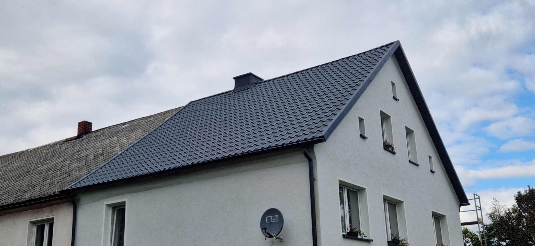 Dom z nowym, grafitowym dachem z blachodachówki trapezowej, widoczne rynny i komin. Część dachu po lewej stronie budynku jest jeszcze stara.