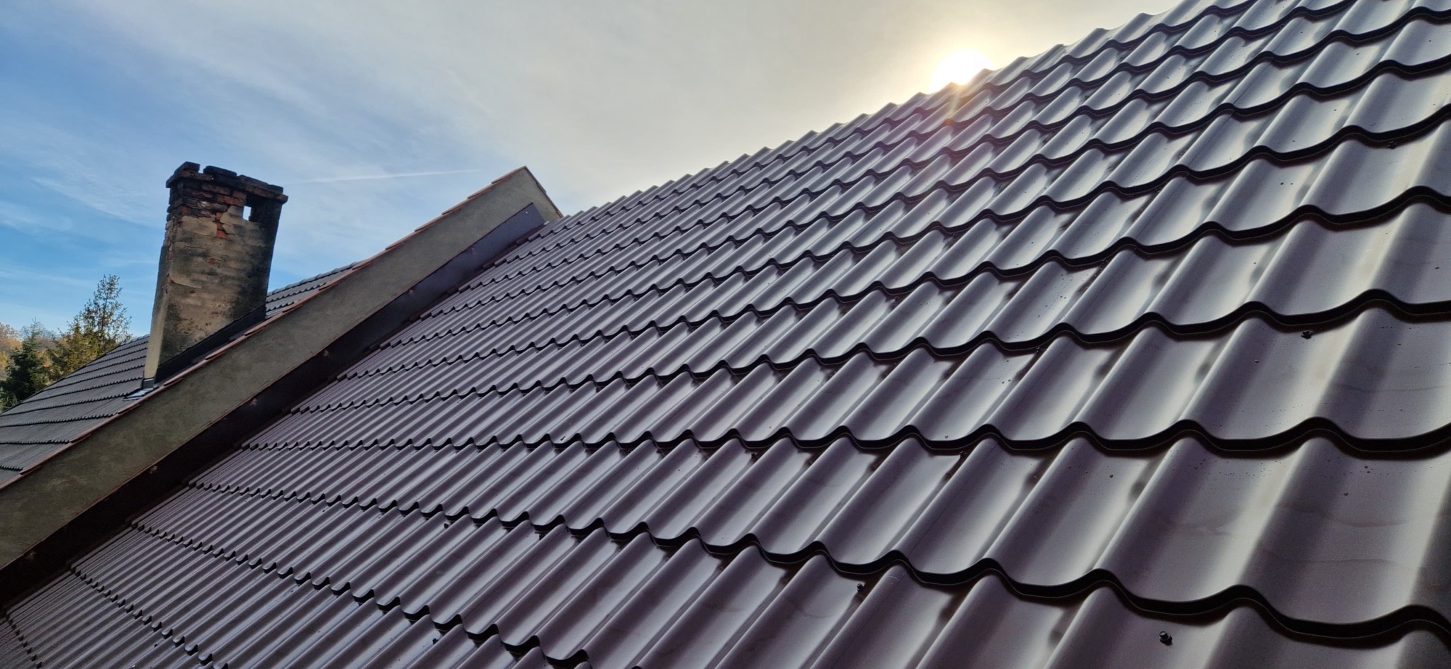 Błyszczący, brązowy dach z blachodachówki w promieniach słońca, z widocznym kominem w tle. Ujęcie z dołu, podkreślające regularność wzoru pokrycia.
