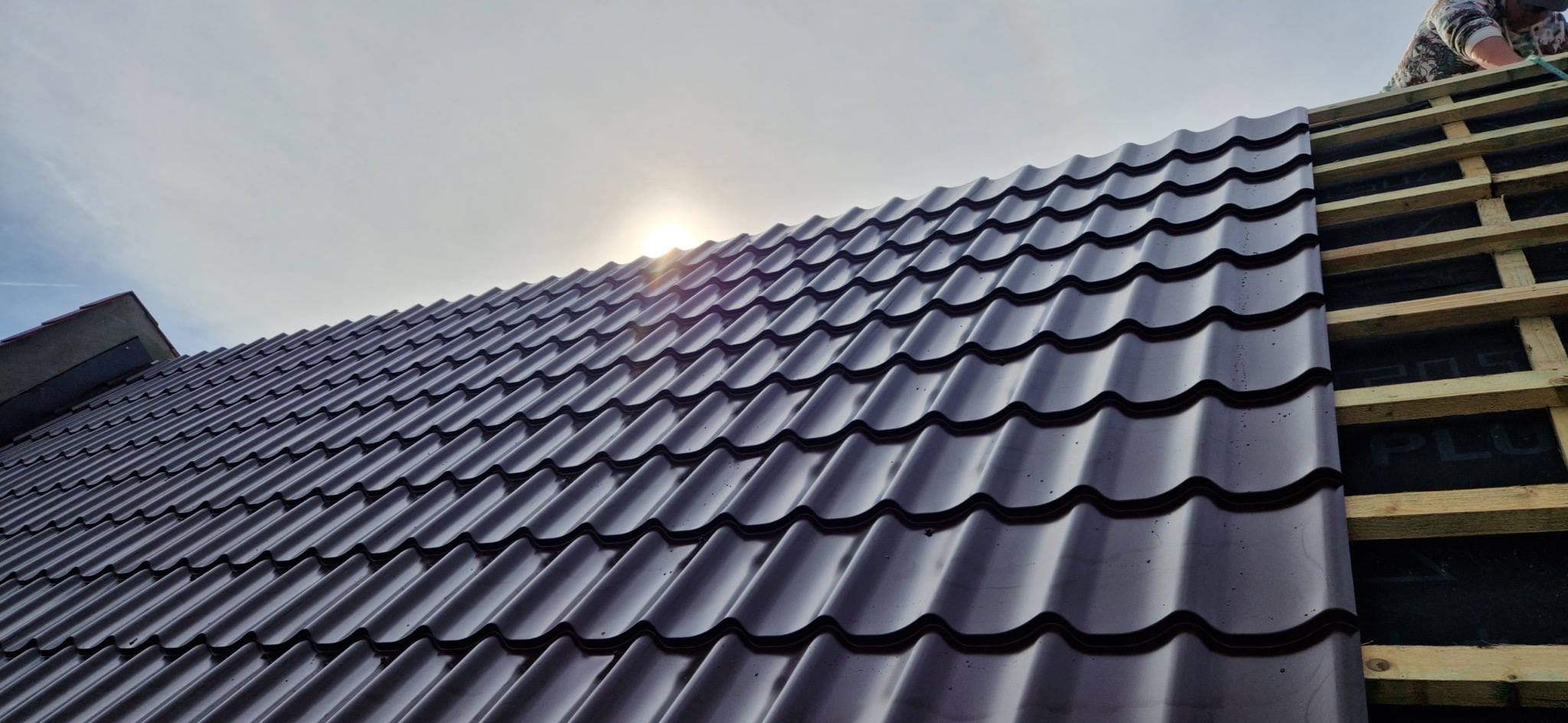 Montaż ciemnobrązowej blachodachówki na łatach dachowych, widok z dołu pod słońce. Częściowo pokryty dach, widoczna konstrukcja.