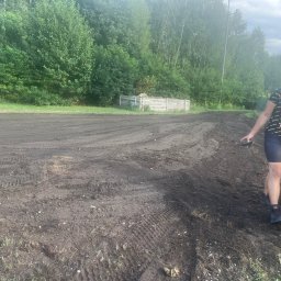 PKUBI-BAU Piotr Kubiciel - Przygotowany teren pod zasiew trawy, widoczne ślady opon i osoba rozprowadzająca nawóz. W tle drzewa i drewniana konstrukcja.