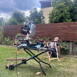 PKUBI-BAU Piotr Kubiciel - Montaż drewnianego ogrodzenia horyzontalnego w ogrodzie. Widoczna piła stołowa, narzędzia i pracownik przytwierdzający deski do słupków. Tło: zieleń i pochmurne niebo.