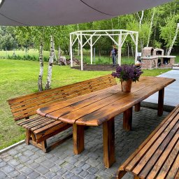 PKUBI-BAU Piotr Kubiciel - Rustykalny stół z ławkami na kamiennym patio, w tle biała pergola w budowie, grill z cegły i pracownik z łopatą. Całość w otoczeniu zieleni i drzew.