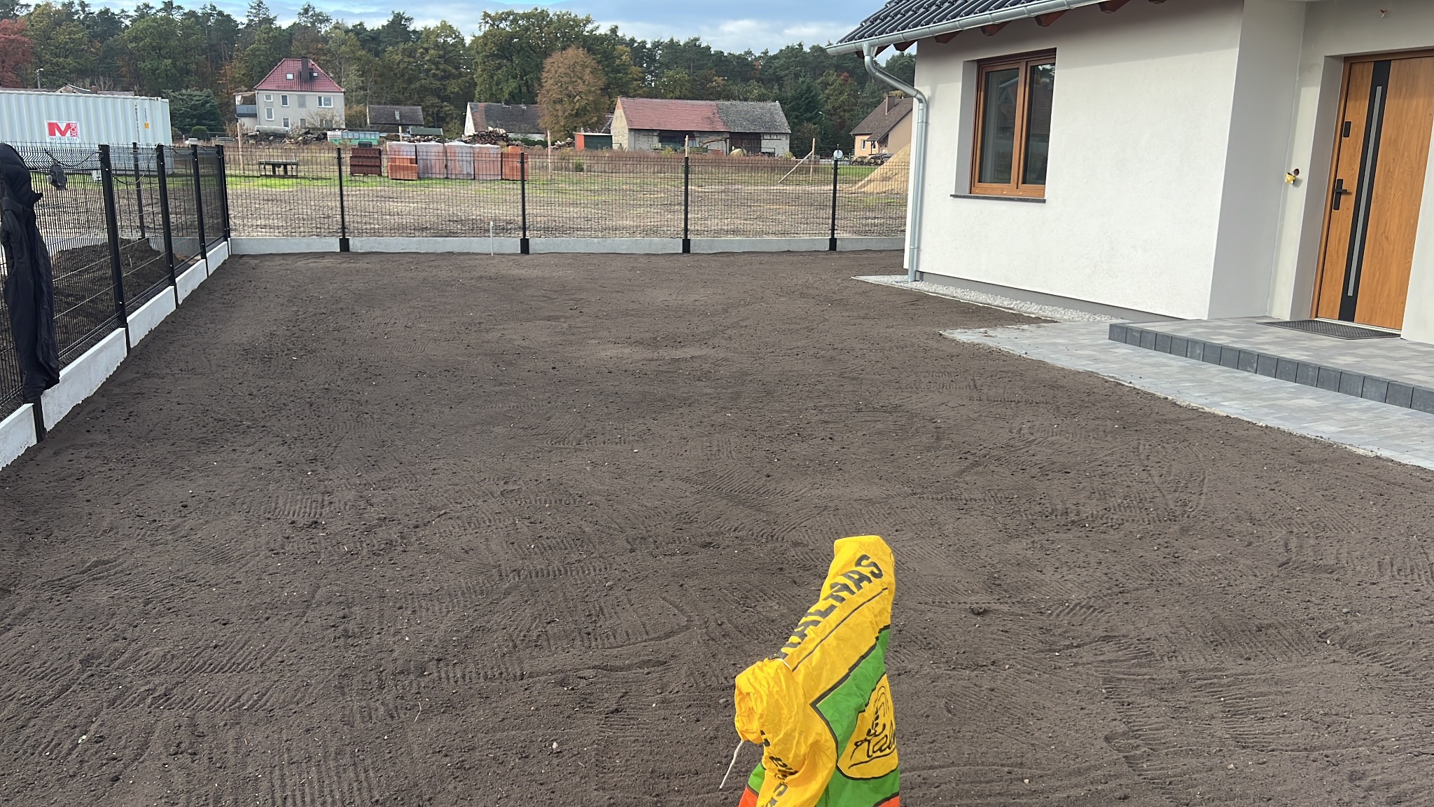 Przygotowany teren pod trawnik z siatką ogrodzeniową na betonowej podmurówce, obok domu z drewnianymi drzwiami i oknem. Widoczny worek z nawozem.