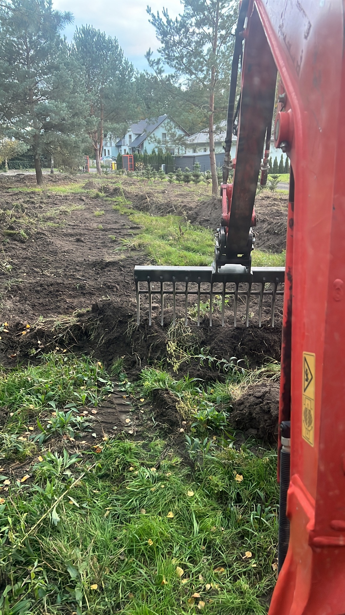 Czerwony traktor z grabiami wyrównuje teren pod budowę na działce z zieloną trawą i drzewami w tle. Widok z bliska na narzędzie robocze.