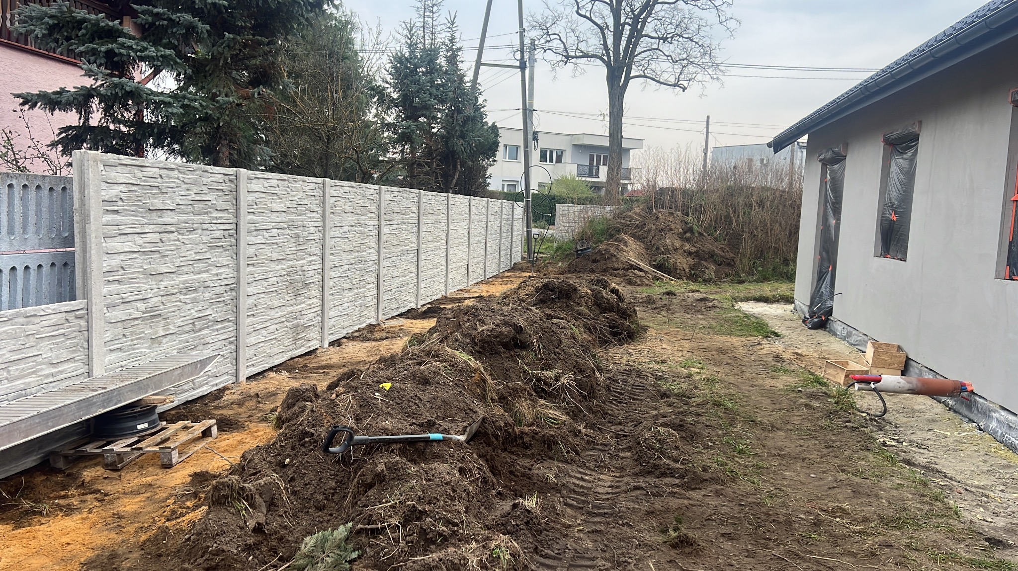 Długi, szary, betonowy płot z fakturą kamienia, w trakcie montażu. Widoczny wykop i narzędzia. Tło: dom, drzewa. Ujęcie z poziomu gruntu.