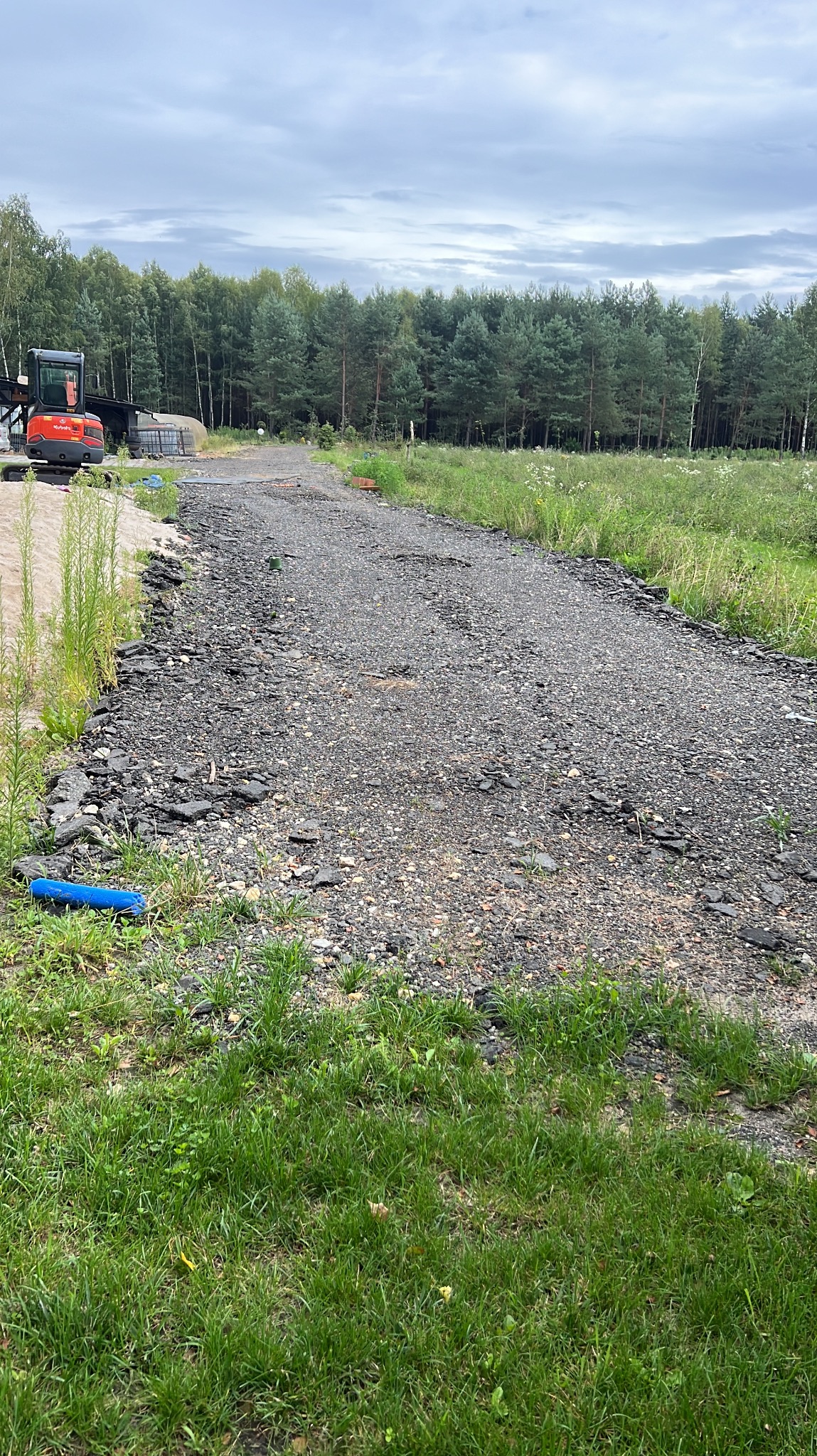 Utwardzona droga gruntowa z widocznym fragmentem niebieskiej rury i koparką w tle, otoczona zielenią i lasem w pochmurny dzień.
