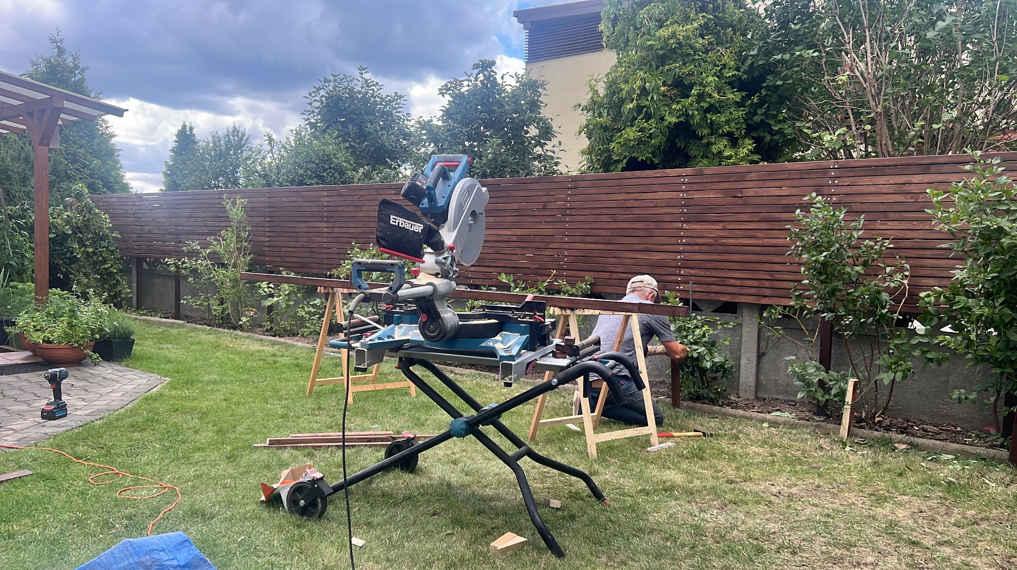 Montaż drewnianego ogrodzenia horyzontalnego w ogrodzie. Widoczna piła stołowa, narzędzia i pracownik przytwierdzający deski do słupków. Tło: zieleń i pochmurne niebo.