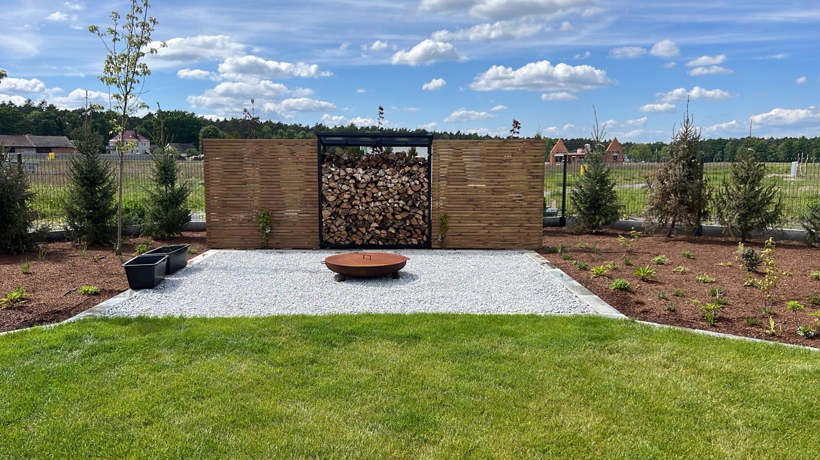 Nowoczesny ogród z drewnianym ogrodzeniem, miejscem na drewno, paleniskiem na żwirze i świeżo posadzoną zielenią. Widok na zadbany trawnik i błękitne niebo.
