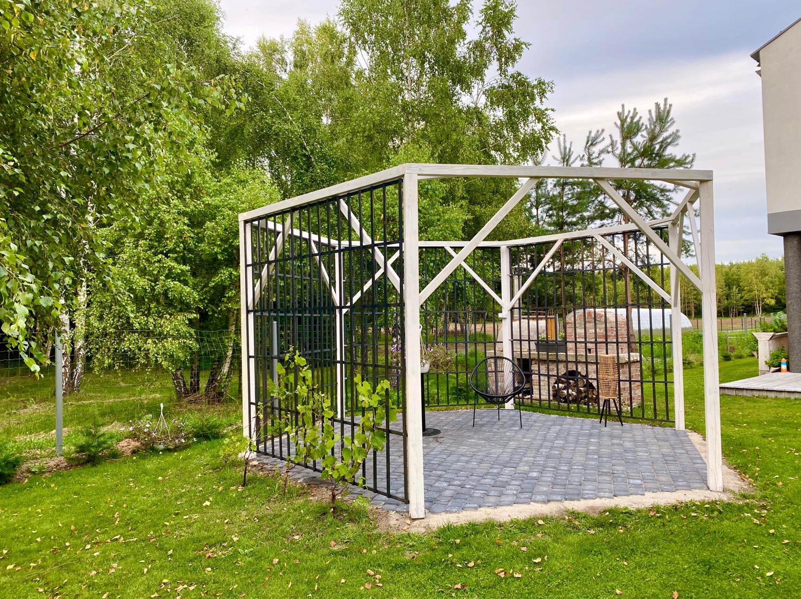 Nowoczesna pergola z czarnymi metalowymi prętami i białymi ramami, z roślinami pnącymi, na tle zielonego trawnika i drzew. Wewnątrz meble ogrodowe.