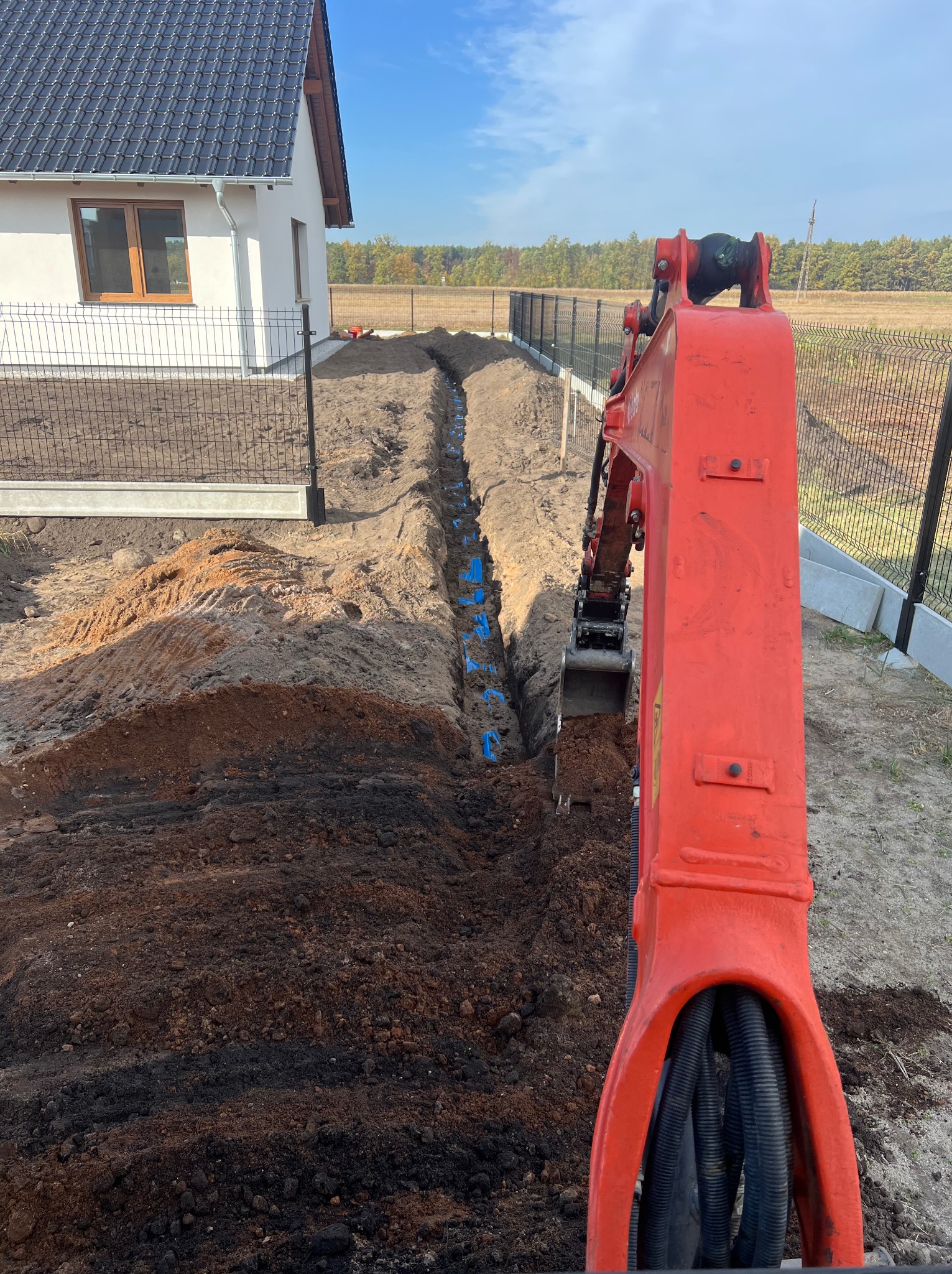Czerwona koparka w trakcie wykopu wąskiego rowu pod instalacje, widoczne niebieskie rury. W tle dom z ciemną dachówką i ogrodzenie z siatki.