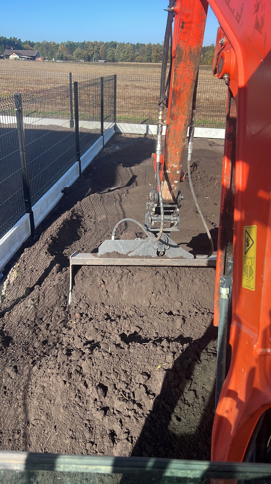 Czerwona koparka wyrównuje ziemię wzdłuż betonowej podmurówki pod ogrodzenie panelowe na tle pola i lasu. Widok z kabiny operatora.