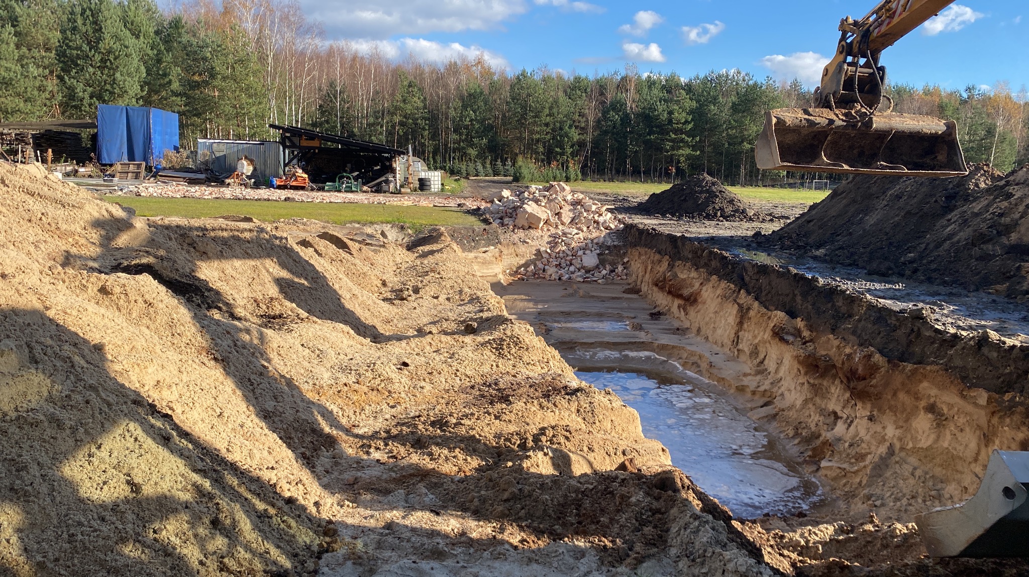 Szeroki wykop z wodą na dnie, z hałdą gruzu pośrodku. W tle budynki gospodarcze i las. Żółta koparka częściowo widoczna po prawej stronie kadru.