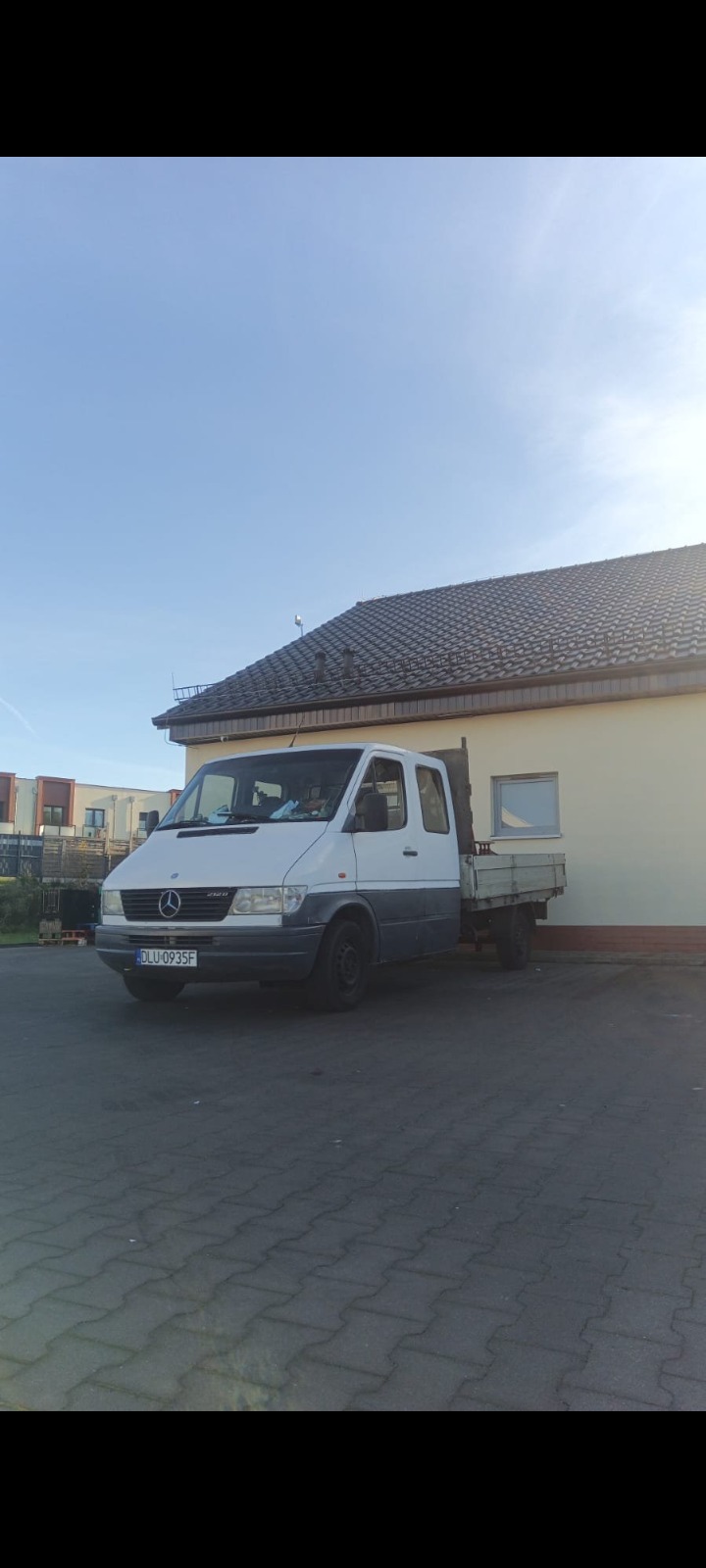 Biały bus Mercedes z otwartą skrzynią ładunkową, zaparkowany na brukowanym placu przed budynkiem z brązowym dachem w słoneczny dzień.