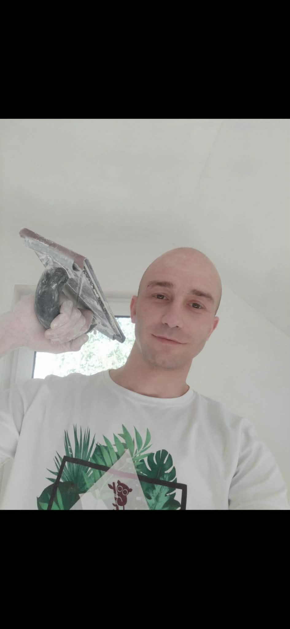 Uśmiechnięty mężczyzna z gładką głową, w białej koszulce z motywem roślinnym, trzyma w rękawicy szpachelkę na tle białej ściany i okna.