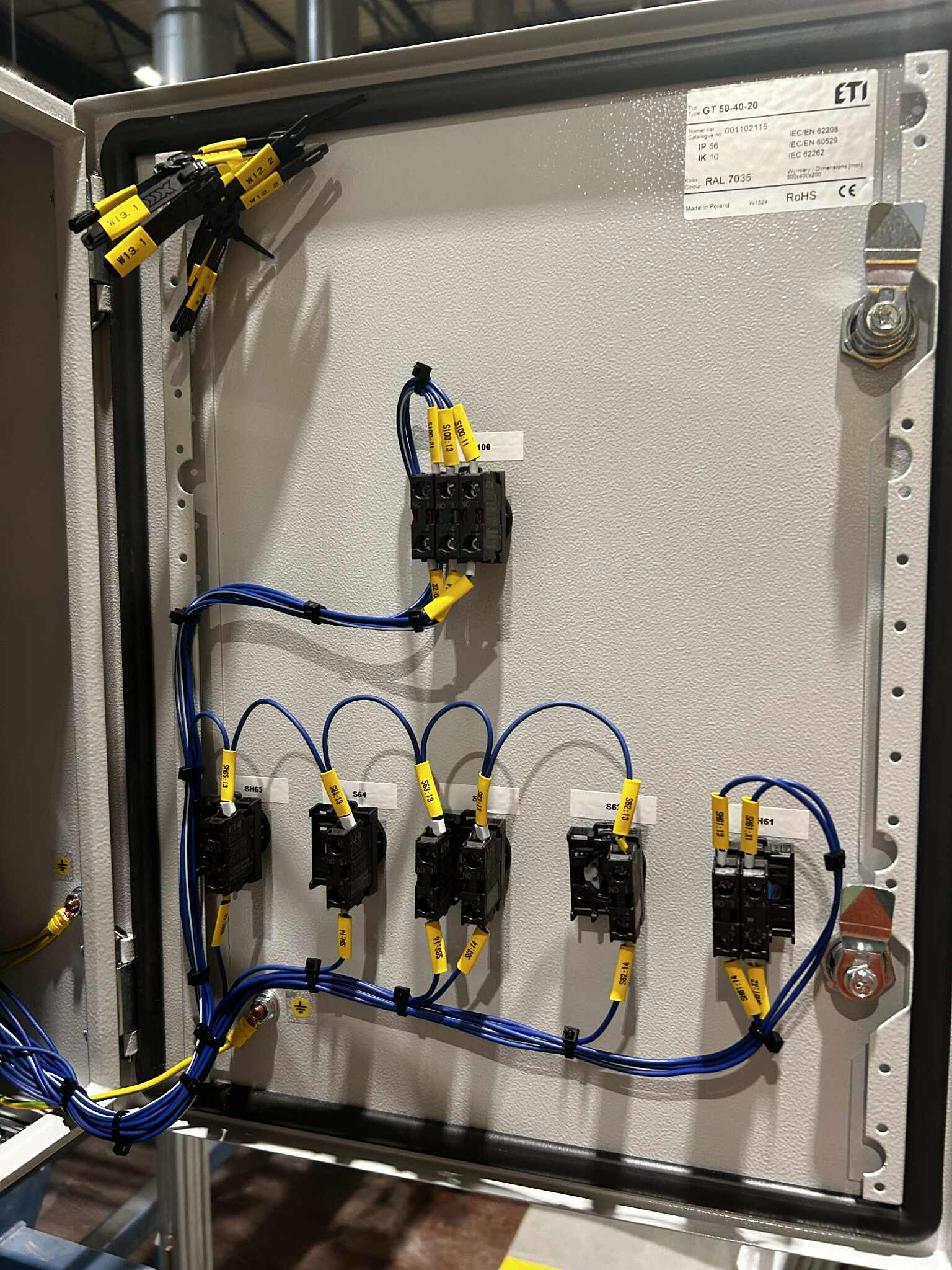 Rozdzielnica elektryczna ETI z okablowaniem oznaczonym żółtymi etykietami, widoczne bezpieczniki i złączki. Instalacja w szarej obudowie z oznaczeniami producenta.