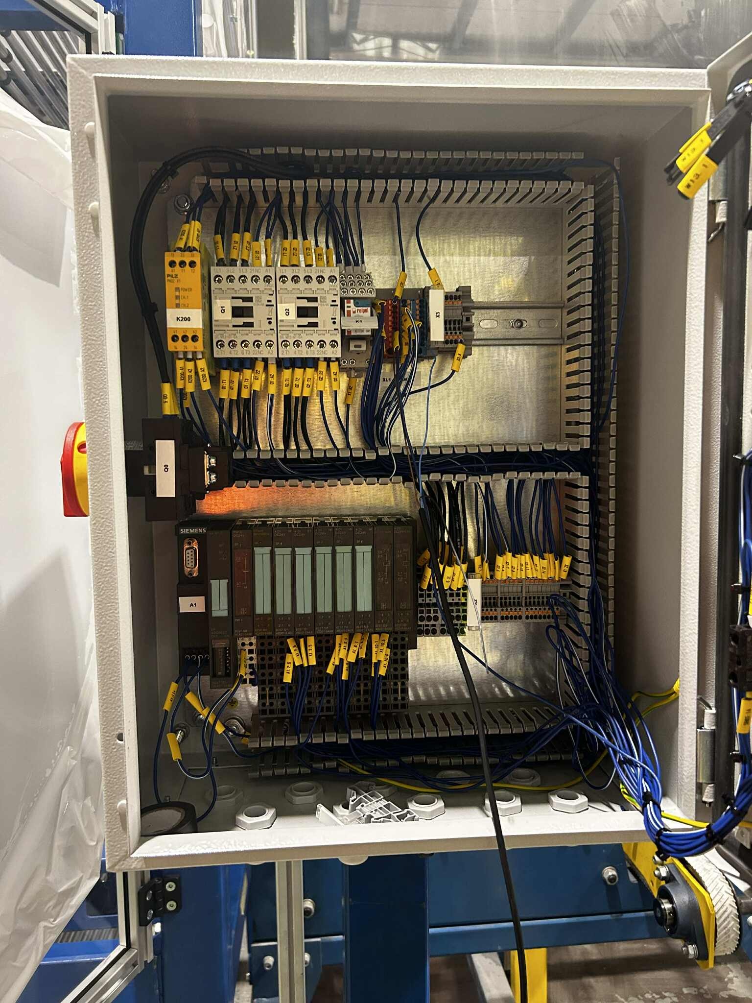 Otwarta szafa sterownicza z modułami Siemens, okablowaniem oznaczonym żółtymi etykietami i elementami zabezpieczającymi Pilz K200. Widoczne złącza i listwy zaciskowe.