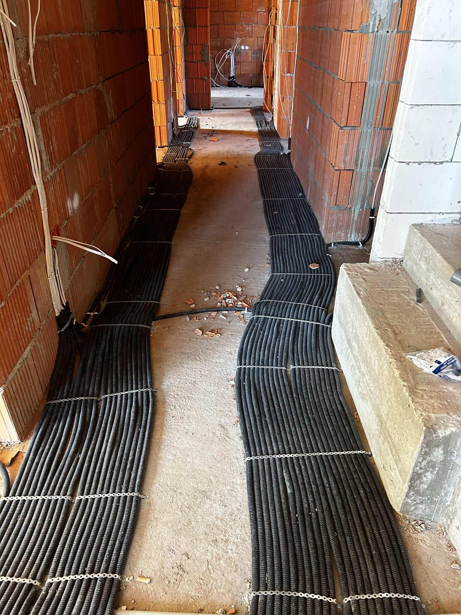 Korytarz w trakcie budowy z ułożonymi kablami elektrycznymi w czarnych peszlach, przymocowanymi metalowymi spinkami do podłoża. Widoczne ściany z czerwonej cegły.