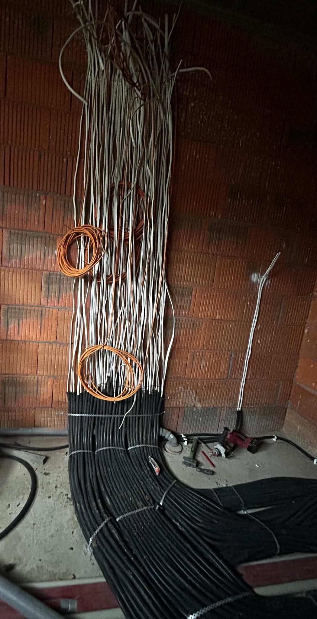 Wiązki kabli elektrycznych w kolorach białym, czarnym i pomarańczowym, ułożone wzdłuż ceglanej ściany, z widocznymi narzędziami pracy na podłodze.