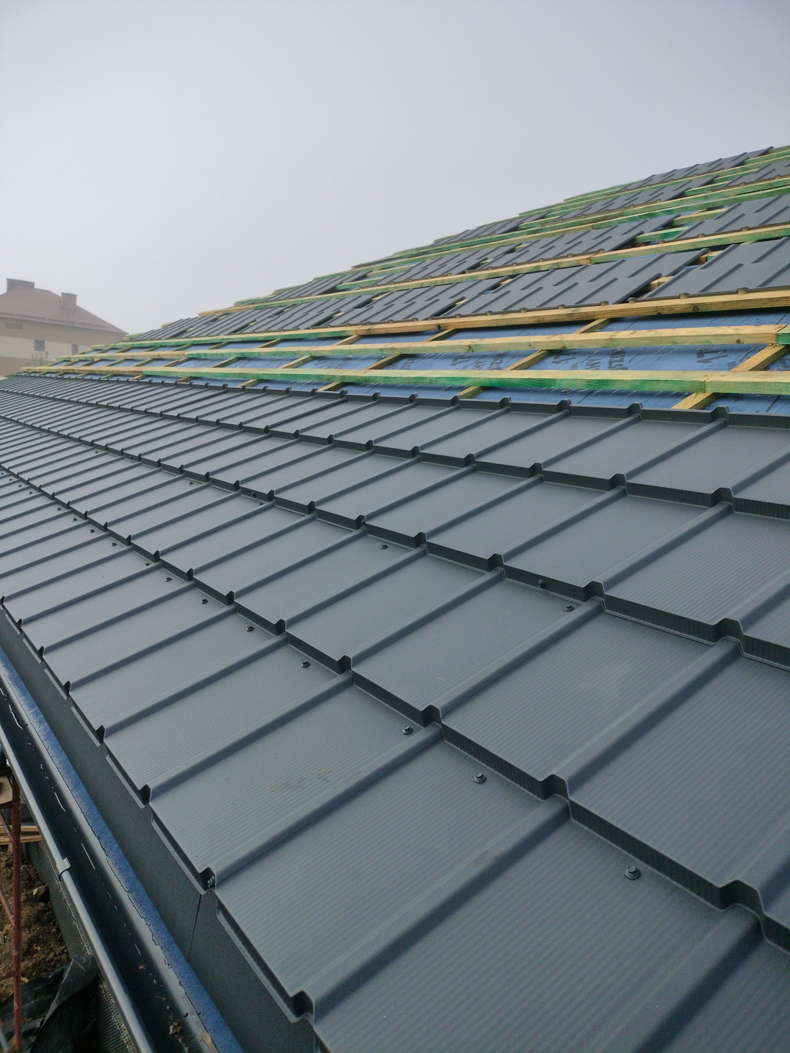 Szary dach z blachy trapezowej w trakcie montażu, widoczne łaty i membrana. W tle zarys budynku w porannej mgle.