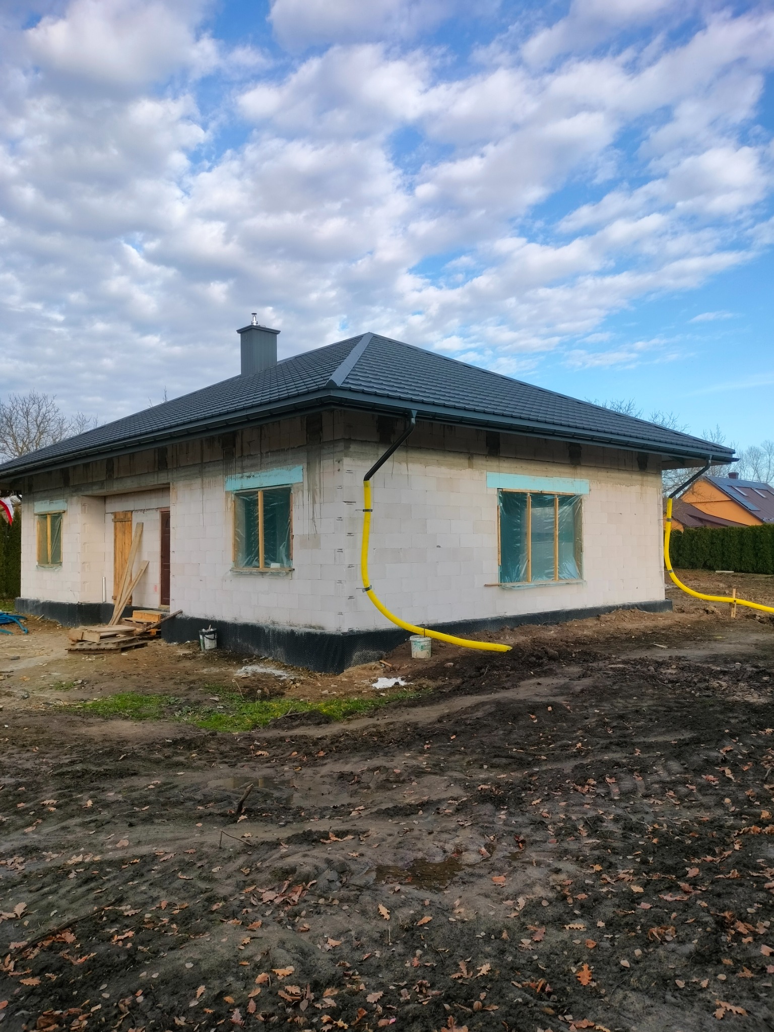 Dom w trakcie budowy z ciemnym dachem i rynnami. Widoczne okna zabezpieczone folią, teren wokół błotnisty. Niebo z chmurami w tle.