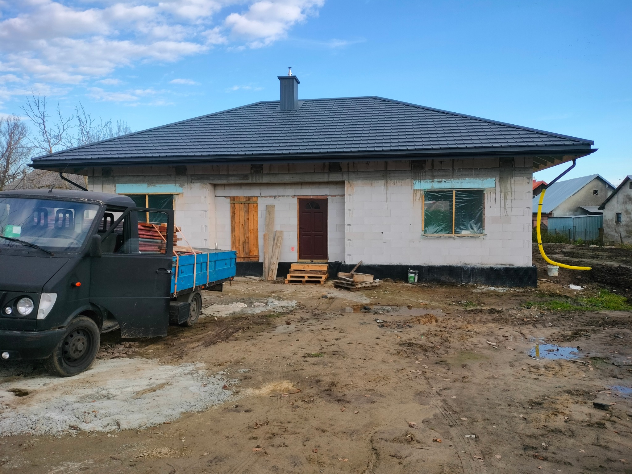 Dom w trakcie budowy z nowym, grafitowym dachem z blachodachówki. Widoczny fragment samochodu dostawczego i teren budowy.
