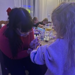 Mimifun - Dorosła w czerwonym swetrze i okularach maluje wzór na przedramieniu blondwłosej dziewczynki. W tle inne dzieci i dzbanek z wodą z cytryną. Scena oświetlona mieszanym światłem.