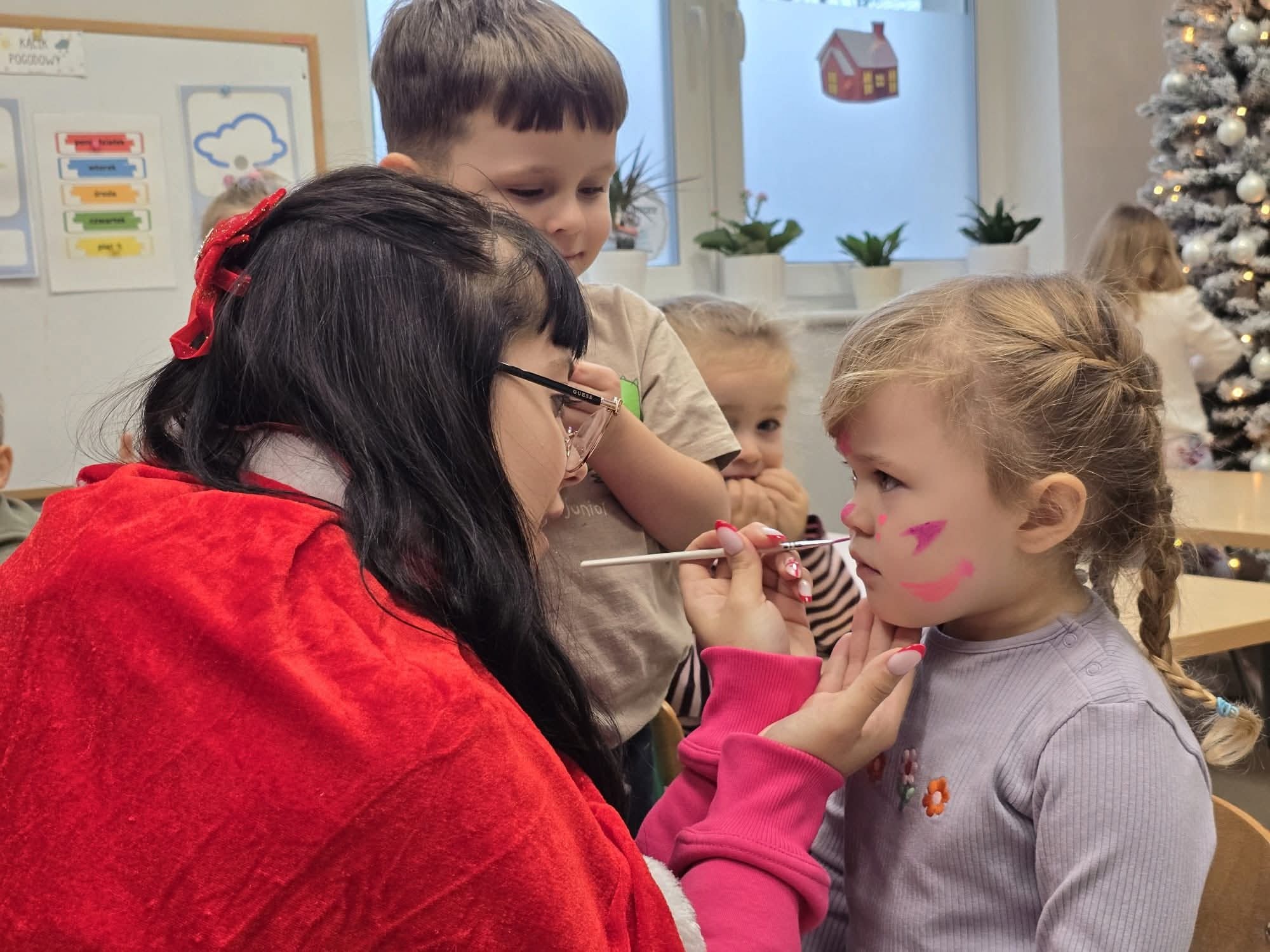 Animator maluje twarz dziewczynce w przedszkolu, używając pędzla i różowej farby. W tle dzieci i choinka. Kadr z bliska, ciepłe oświetlenie.