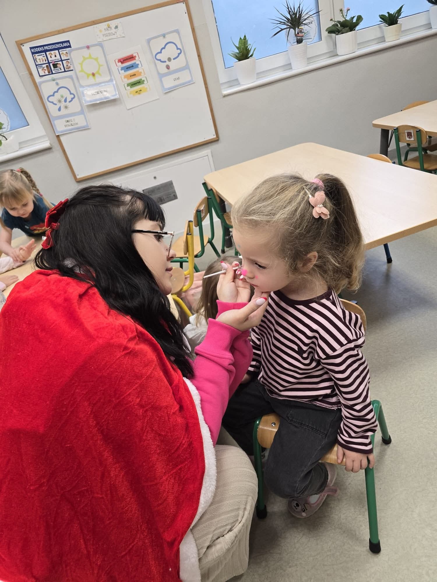 Animator maluje twarz dziewczynki w przedszkolu. Kobieta w czerwonym stroju, dziecko w pasiastym ubraniu. W tle tablica z pogodą i rośliny doniczkowe.