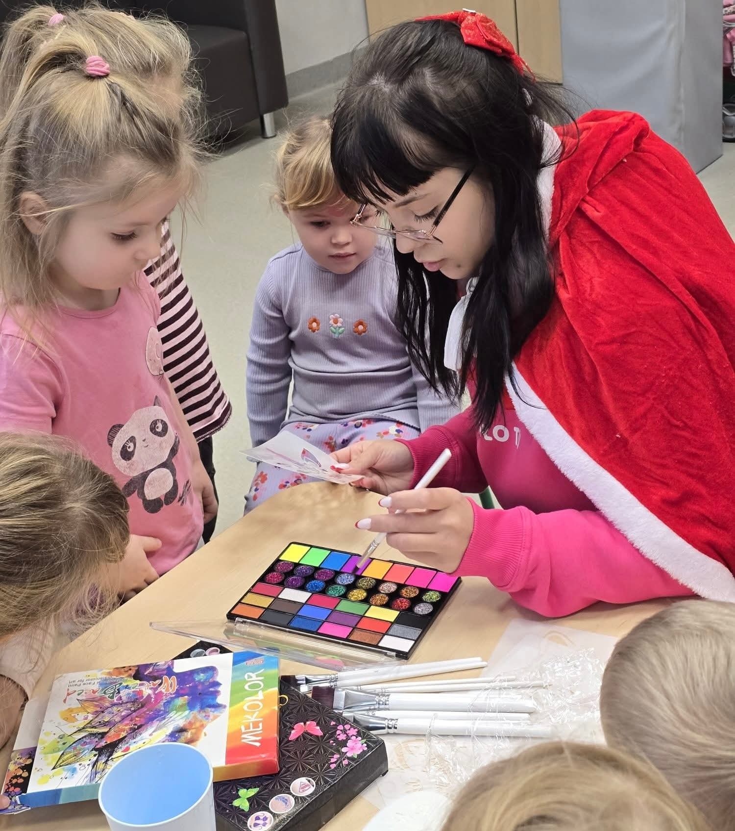 Animator w czerwonym płaszczu maluje twarze dzieciom w przedszkolu. Paleta kolorów, pędzle i wzorniki na stole. Dziewczynki z zaciekawieniem obserwują.