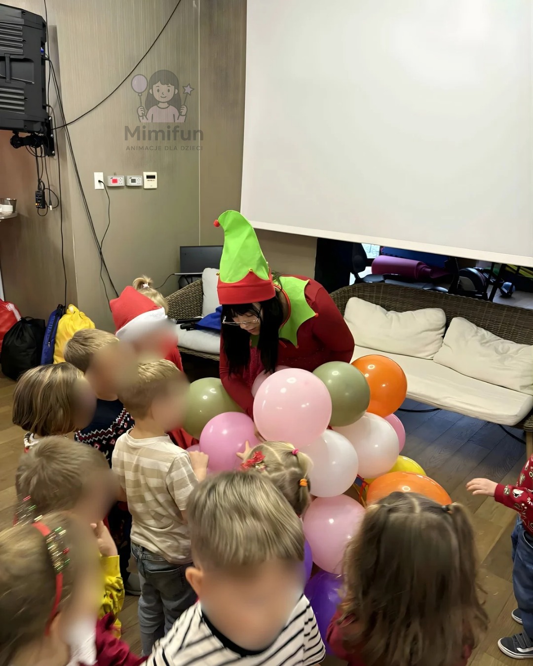 Animator w stroju elfa wręcza balony grupie dzieci podczas animacji w pomieszczeniu. Widoczny sprzęt nagłośnieniowy i logo firmy animacyjnej.