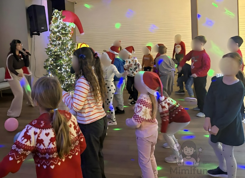 Animator w stroju Mikołaja śpiewa do mikrofonu, dzieci w czapkach Mikołaja tańczą wokół choinki, kolorowe światła dyskotekowe, hotel Szlak Bursztynowy w Jarosławcu.