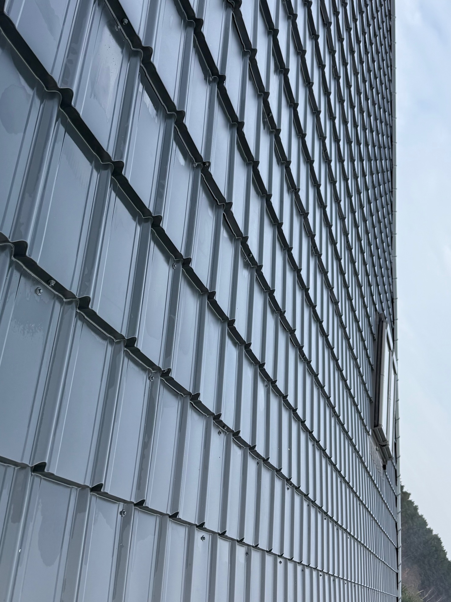 Szary, metalowy dach z widocznymi mocowaniami, sfotografowany pod kątem, z fragmentem okna i drzew w tle. Nowoczesne pokrycie.