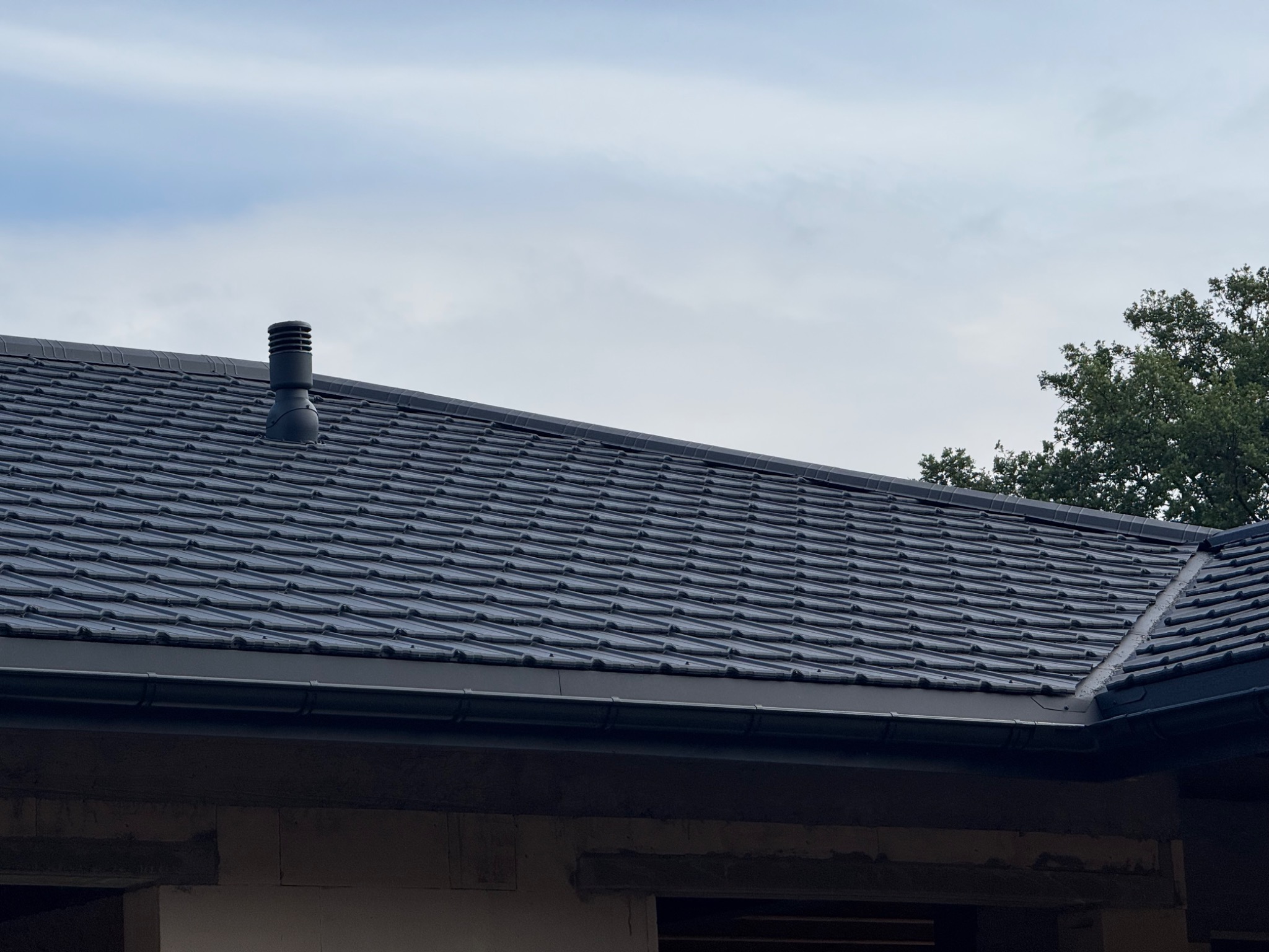 Nowy, grafitowy dach z ceramicznej dachówki karpiówki, z zamontowaną rynną i kominkiem wentylacyjnym, na tle błękitnego nieba i zieleni drzew.