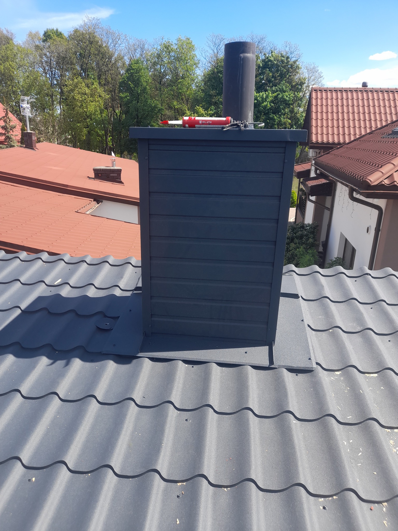 Szary komin wentylacyjny na dachu z blachy trapezowej, widoczny aplikator uszczelniacza. W tle inne dachy i zieleń drzew, błękitne niebo.