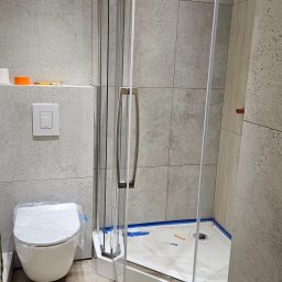 Daniel Poczobut DP Projekt Detal - Nowoczesna łazienka w trakcie wykańczania: kabina prysznicowa z otwartymi drzwiami, toaleta z folią ochronną i szare płytki imitujące beton na ścianach.